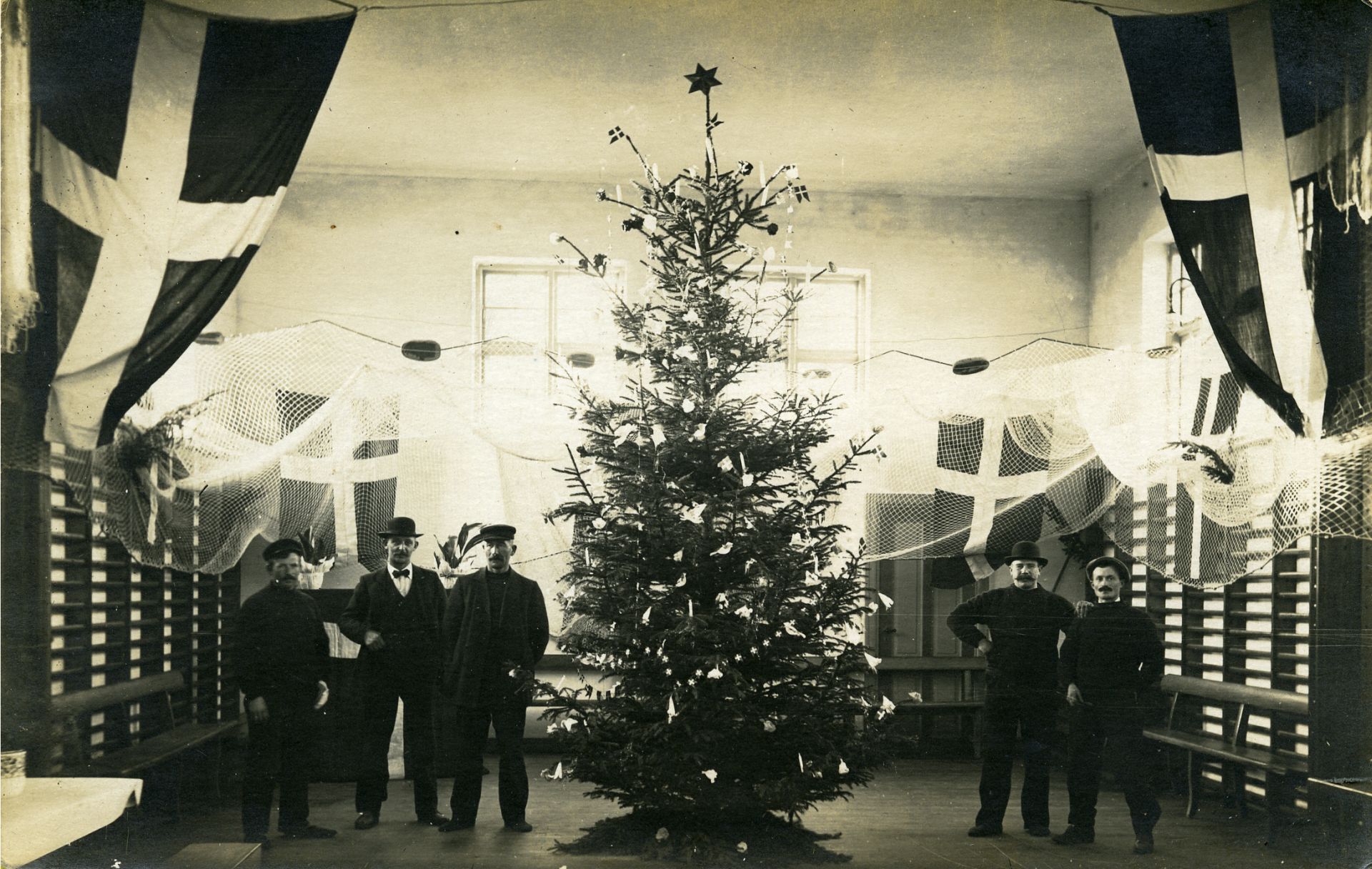 Fem mænd står rundt om et stort juletræ i en gymnastiksal. Der er fiskenet i loftet og fire store dannebrogsflag.