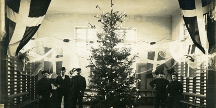 Fem mænd står rundt om et stort juletræ i en gymnastiksal. Der er fiskenet i loftet og fire store dannebrogsflag.